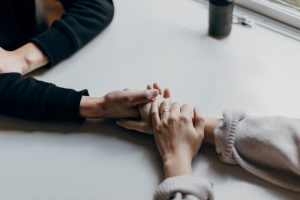 Dos personas se sostienen las manos sobre una mesa junto a una ventana, en un gesto de apoyo emocional.